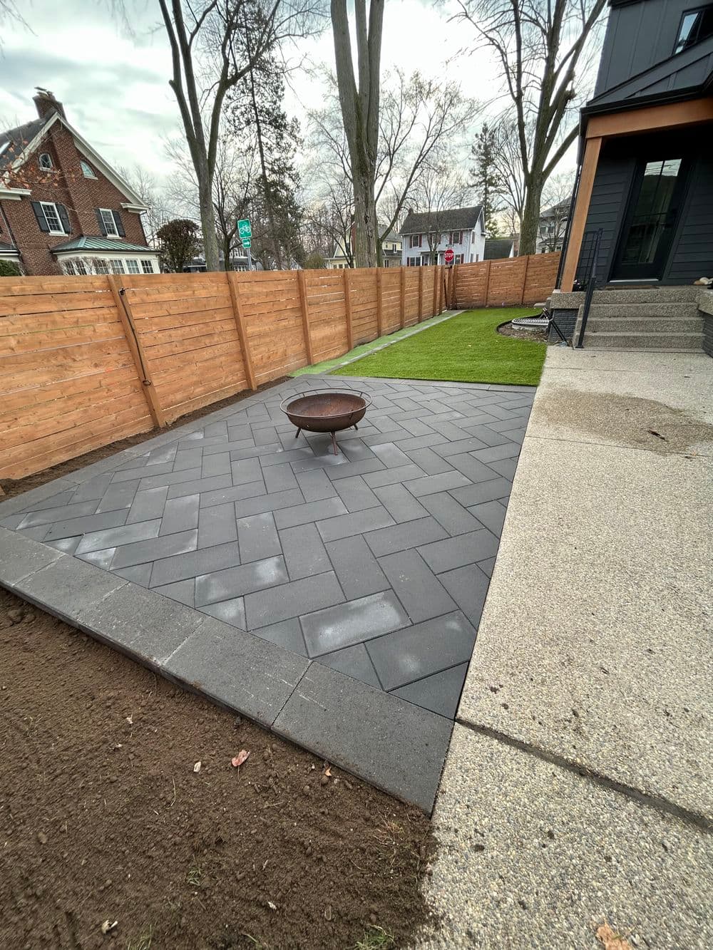 Patio with herringbone pavers and fire pit, surrounded by a wooden fence and lawn.