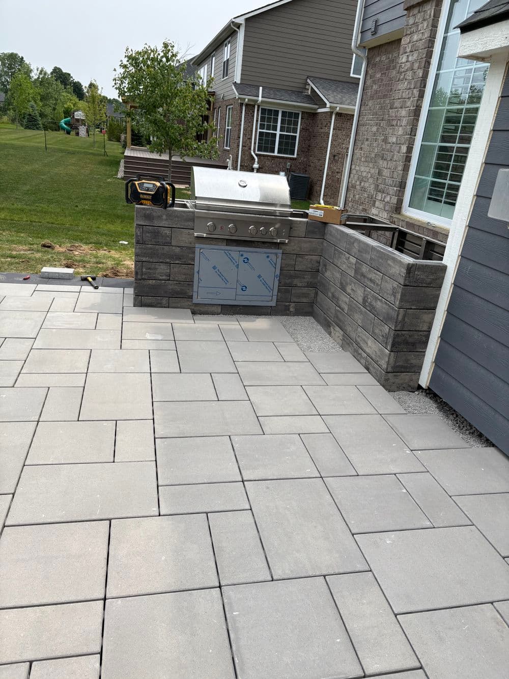 Outdoor kitchen with grill built into stone cabinetry on a paved patio, surrounded by greenery.