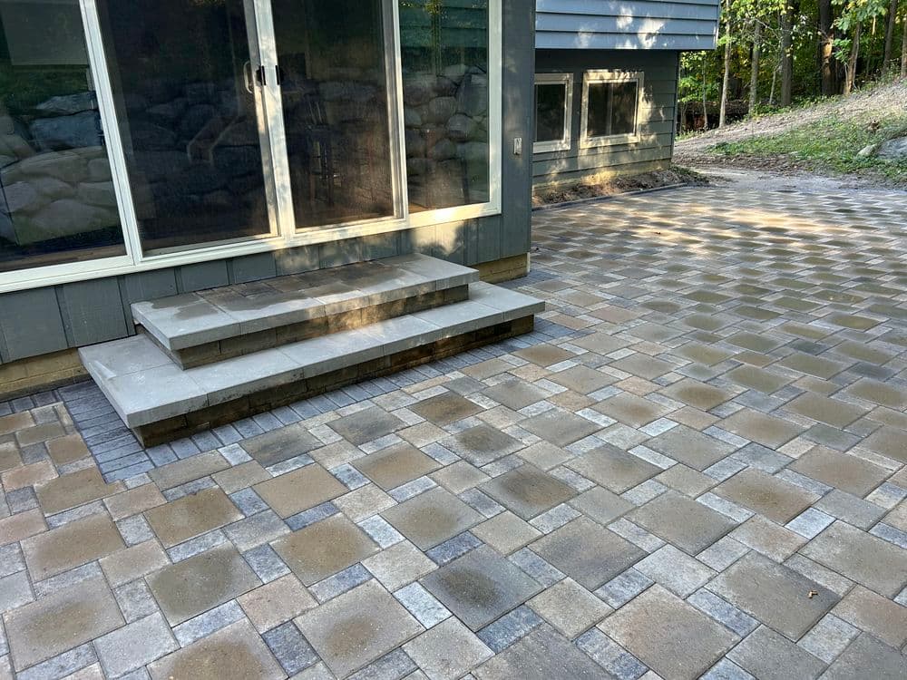 Paver patio with stone steps leading to a home, surrounded by wooded area.