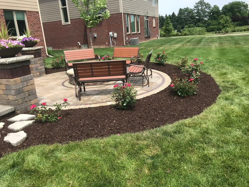 Patio area with wooden chairs, stone wall, and colorful flower beds in a lush green yard.