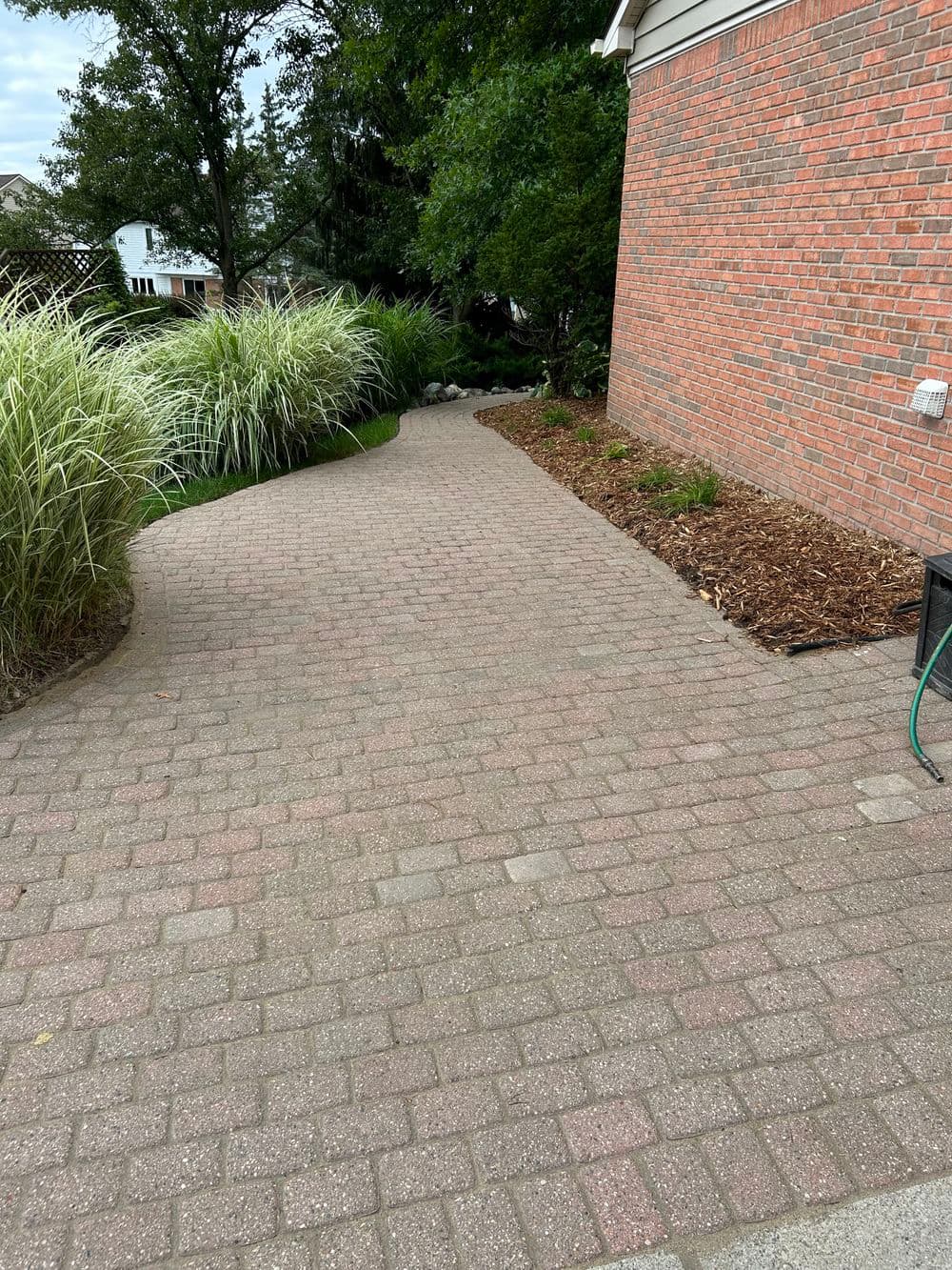Paved walkway beside a brick house, surrounded by greenery and landscaping.