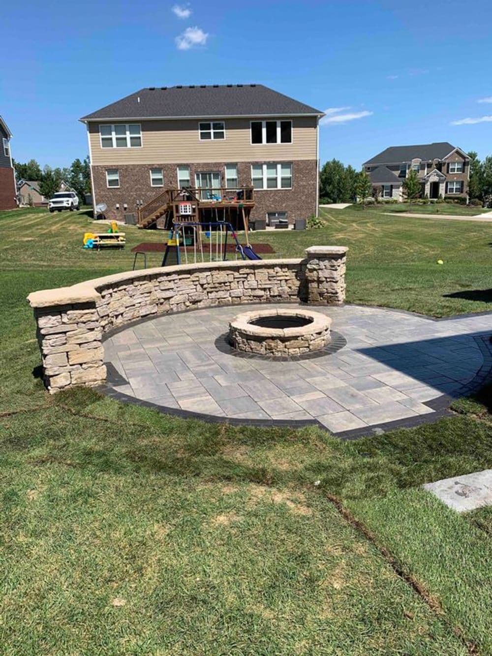 Backyard fire pit surrounded by stone seating and lawn, with houses in the background.