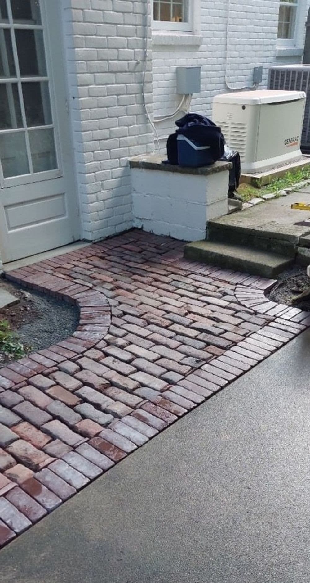 Brick walkway leading to a white house with a porch and utilities visible.