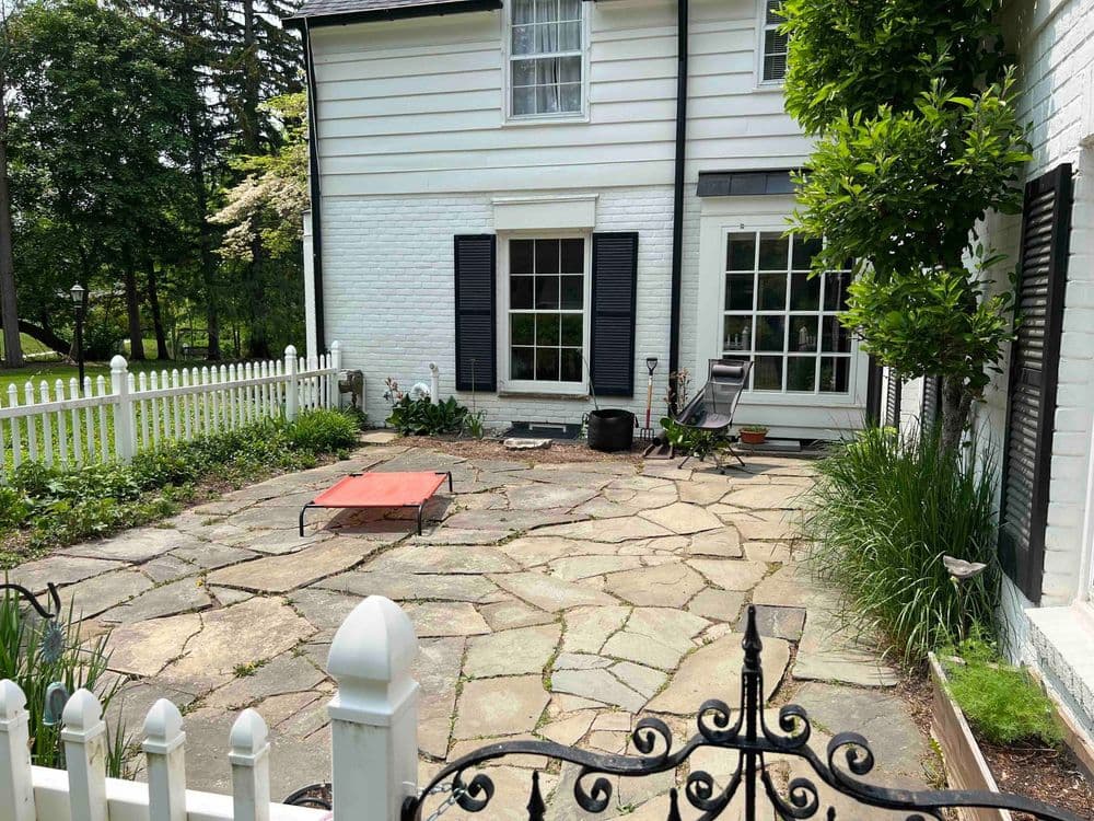 Charming stone patio with white fencing, outdoor seating, and lush greenery in a residential setting.